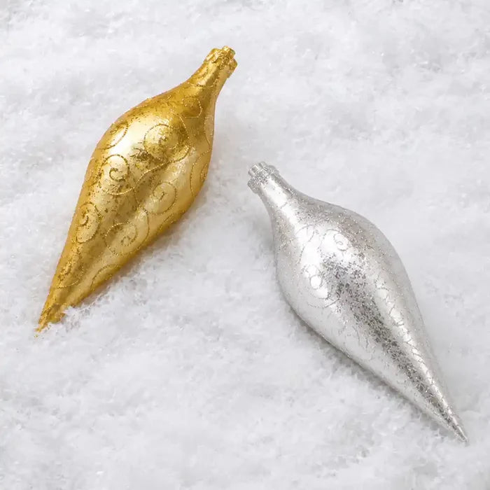 Two decorative Christmas ornaments shaped like teardrop pomegranates, one in metallic gold and one in metallic silver, both featuring embossed scrollwork, resting on a white faux-snow background.