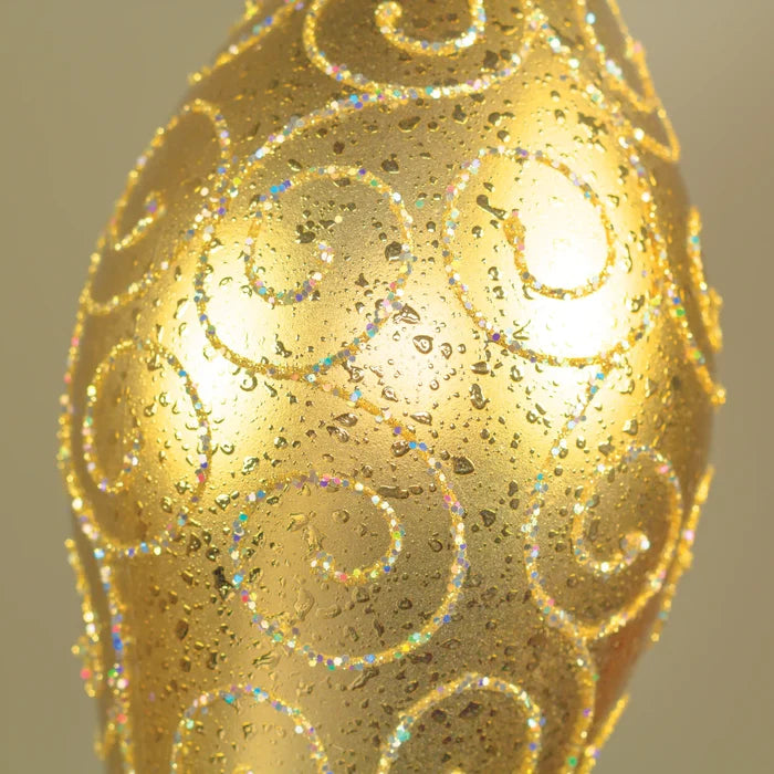 A close-up of a gold sprinkle finial ornament or vase-like object with an iridescent glitter swirl pattern on a soft beige background.